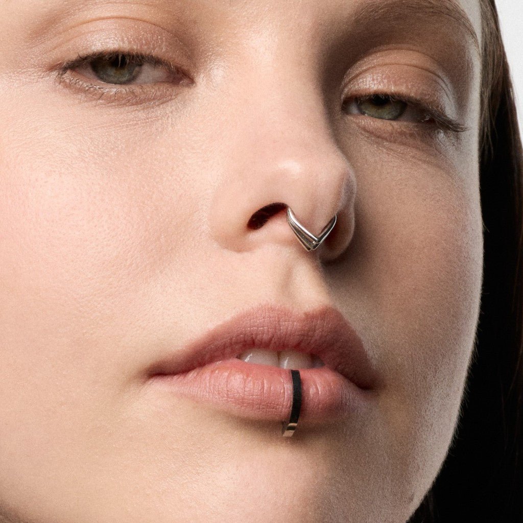 Close-up of a model's face showing a silver chevron nose ring on the septum and a silver curved barbell clicker piercing
.