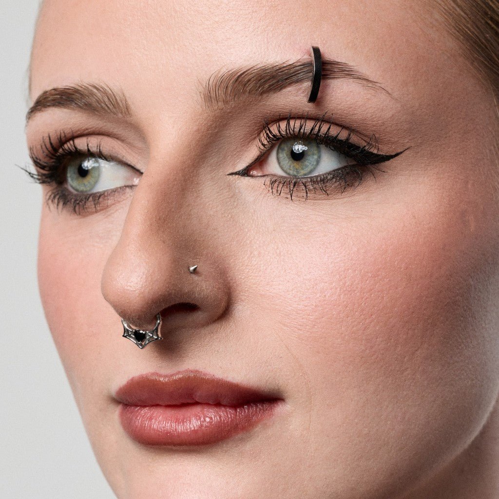 Close-up of a woman's face showing a black eyebrow barbell piercing and a silver nose stud with a black bow detail, against a light gray background.