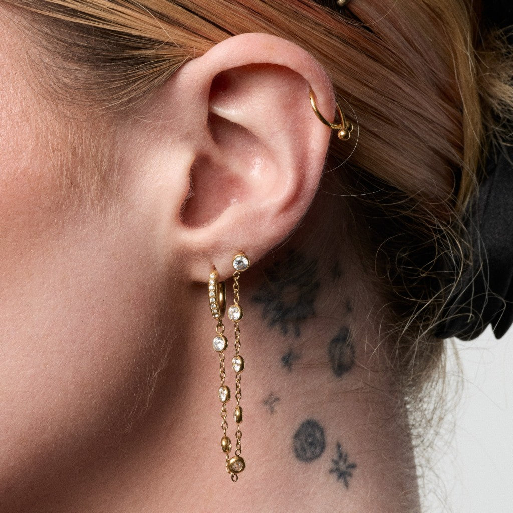 A close-up of a person's ear wearing a pair of gold huggie hoop earrings with dangling chains and clear gemstones, along with a gold hoop in the opposite ear cartilage.