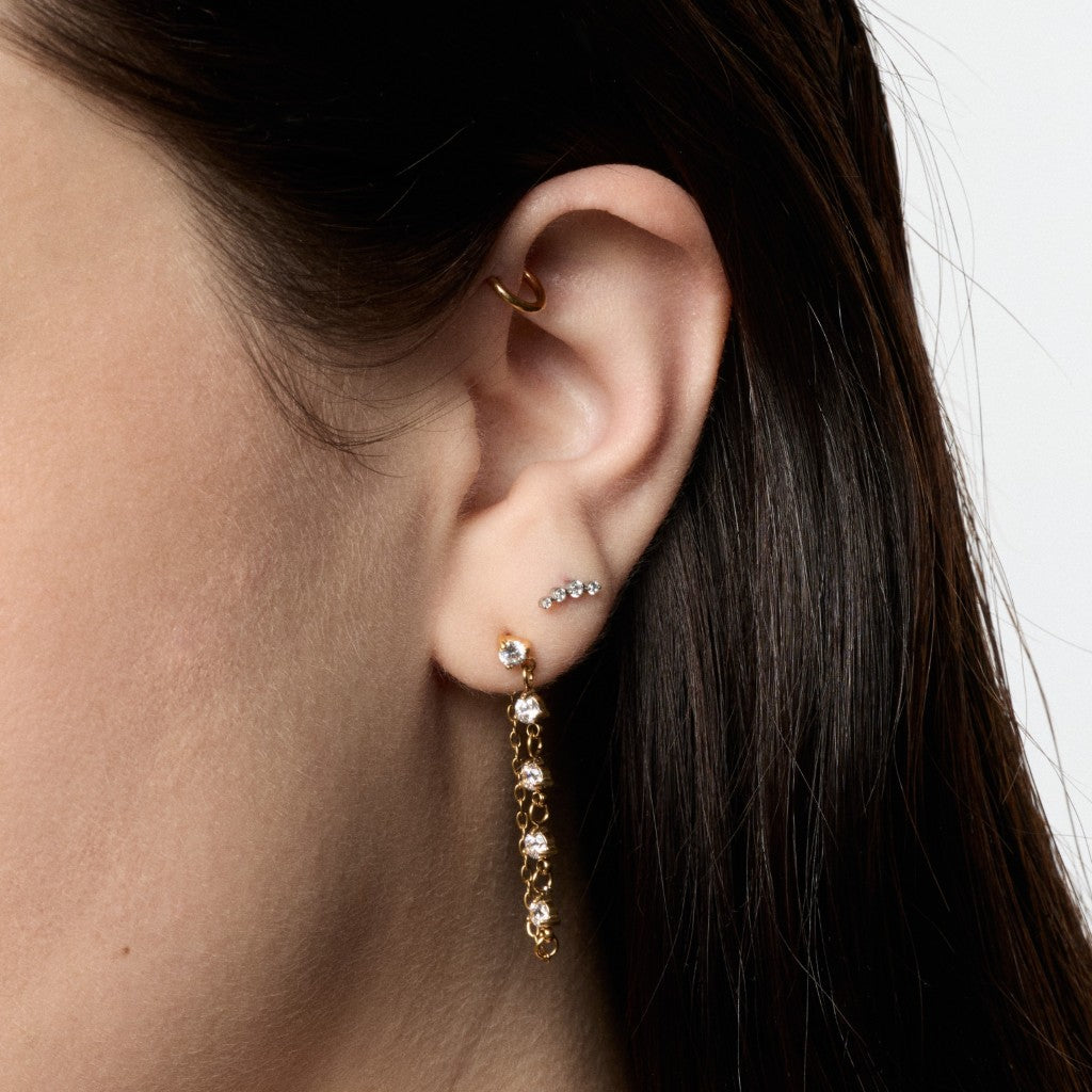 A close-up of a model's ear showcasing a gold dangle hoop earring adorned with clear crystals, alongside other ear piercings.