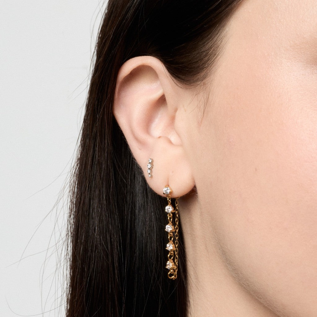 A close-up of a model's ear wearing a gold dangle earring with small clear stones, alongside a smaller matching stud earring, against a light gray background.