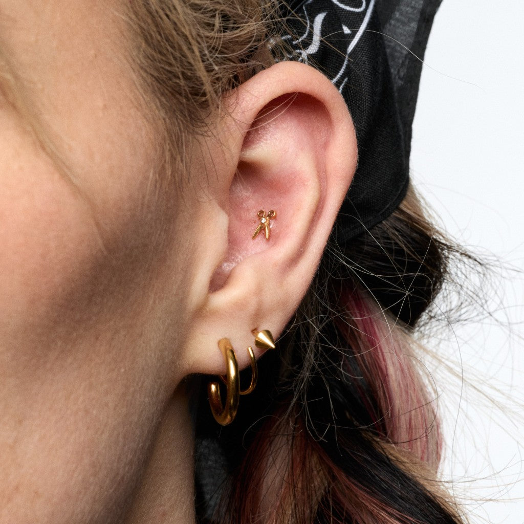 A close-up of a person's ear showcasing gold piercings, including a small bow-shaped stud in the conch, a gold hoop and a small gold flatback in the lobe.