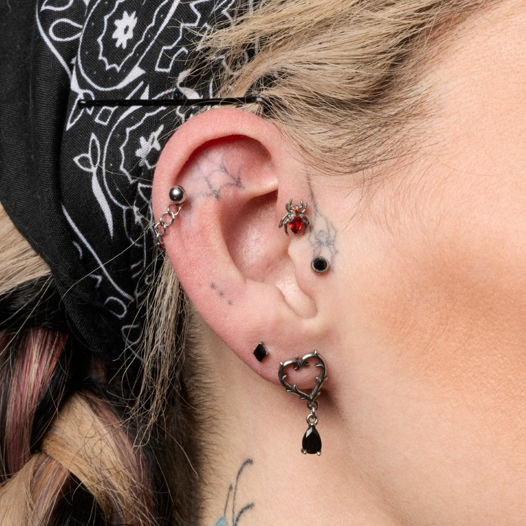 Close-up of a person's ear showcasing multiple piercings, including a silver heart-shaped dangle earring with a black teardrop charm, a silver ear flatback, and other small studs and rings.
