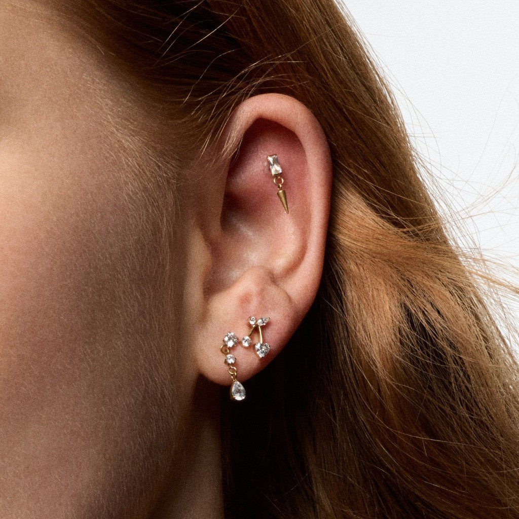 A close-up of a person's ear showcasing multiple gold ear cuffs and studs adorned with clear crystals, set against a clean white background.