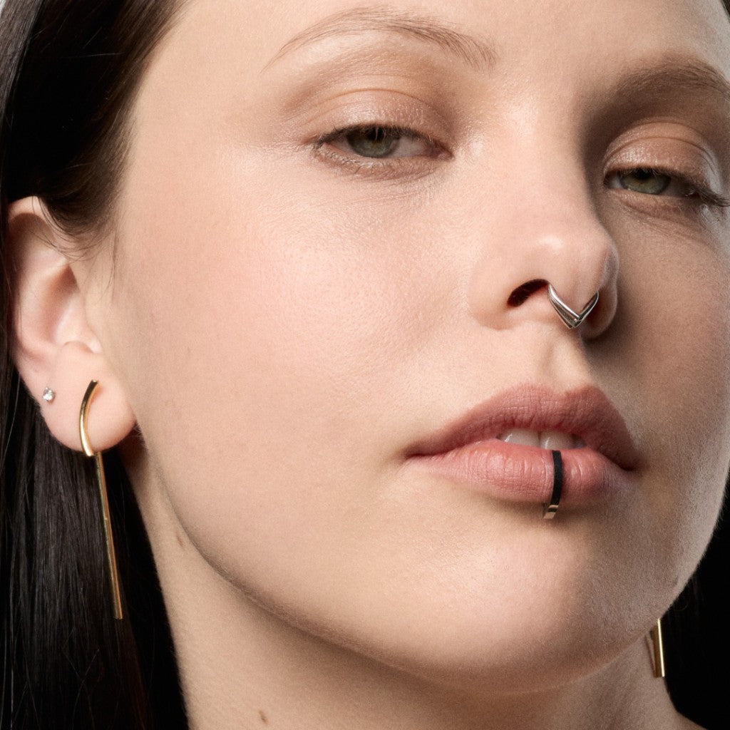 Close-up of a model wearing a gold crescent hoop earring with a dangling barbell drop, a silver V-shaped nose ring, and a curved barbell clicker lip ring.