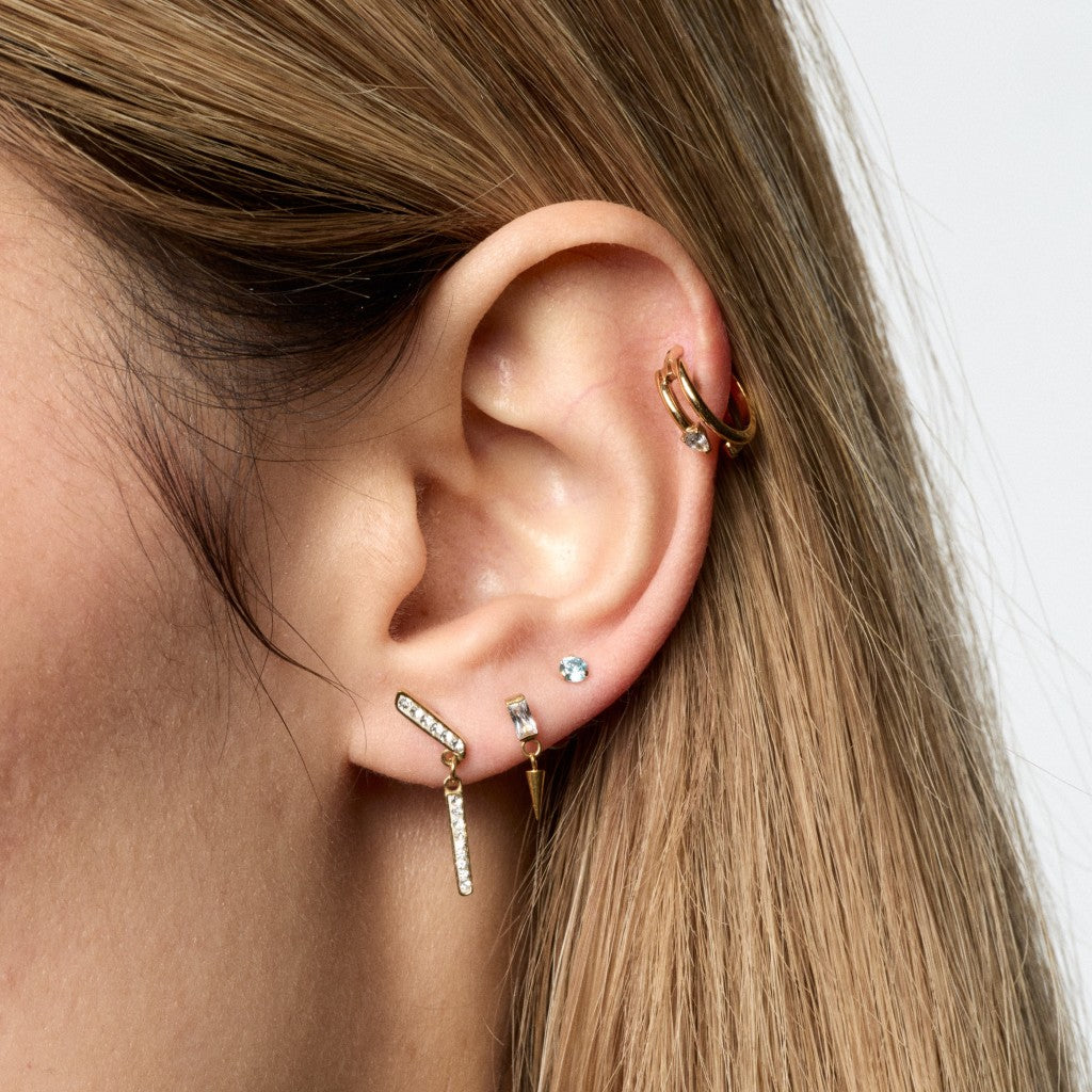 Close-up of a person's ear showcasing multiple gold and silver ear piercings, including a hoop in the helix, a small stud, a barbell with a dangling chevron, and a small vertical stud.