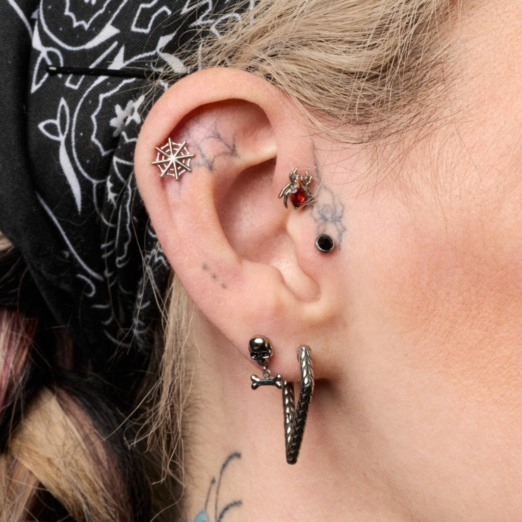 Close-up of a person's ear showcasing multiple gothic and alternative piercings, including a spiderweb flatback, a small skull stud, a snake hoop, and a black bead stud.