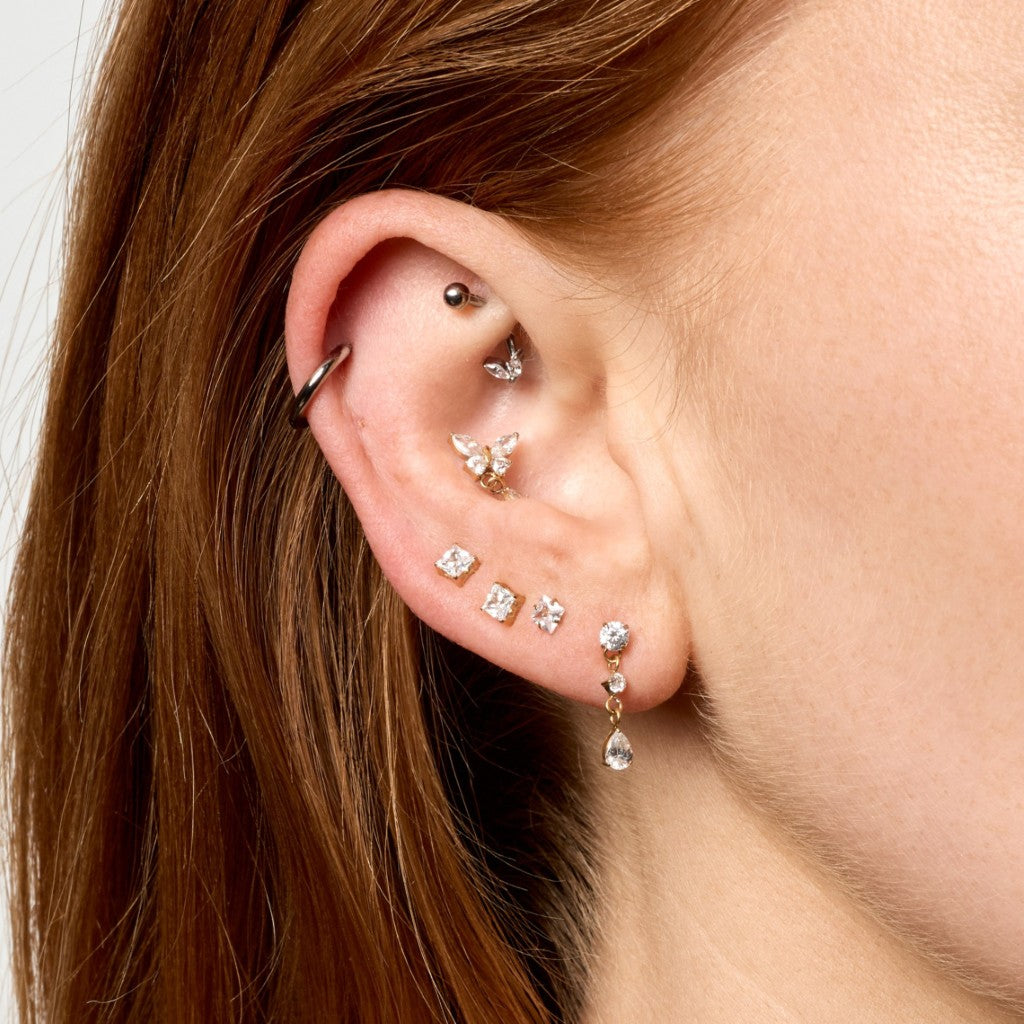 Close-up of a person's ear showcasing multiple gold ear piercings and stud earrings, including a butterfly-shaped stud, a teardrop dangling stud, and small square stud earrings.