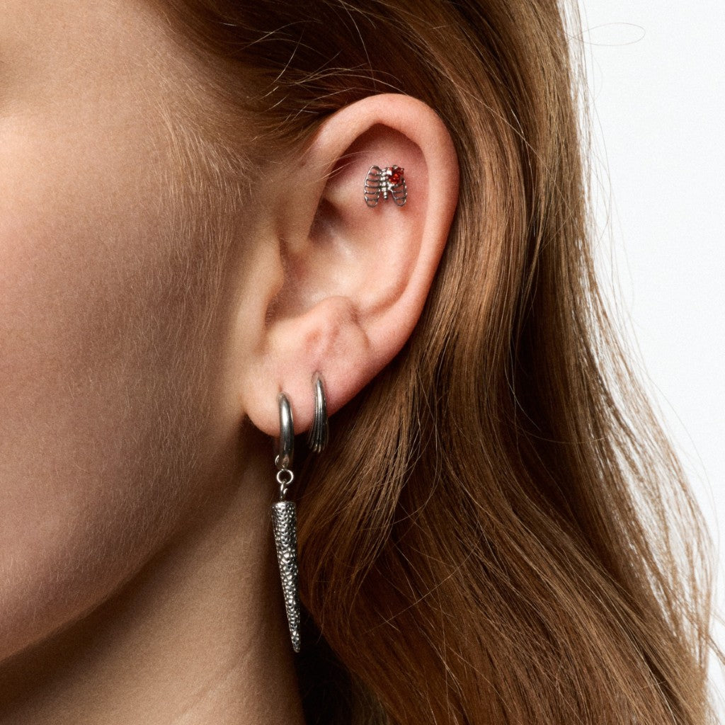 Close-up of a person's ear wearing multiple silver-toned earrings, including a small ribcage-shaped flatback with a red centre and a dangling textured hoop earring.