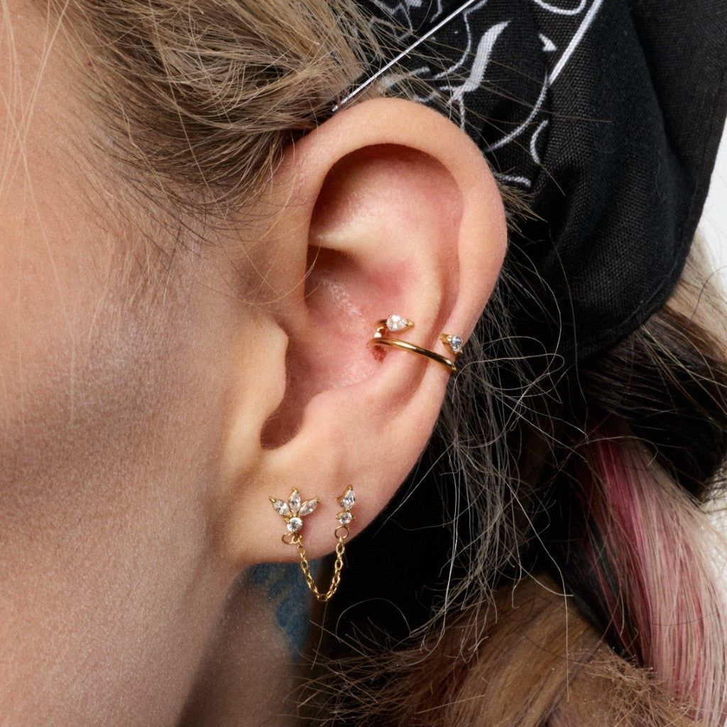 A close-up of a person's ear showcasing multiple gold ear piercings: a curved hoop in the upper lobe, a small marquise-shaped lobe stud with tiny clear stones and a dangling chain, and a flat gold flatback in the lobe.