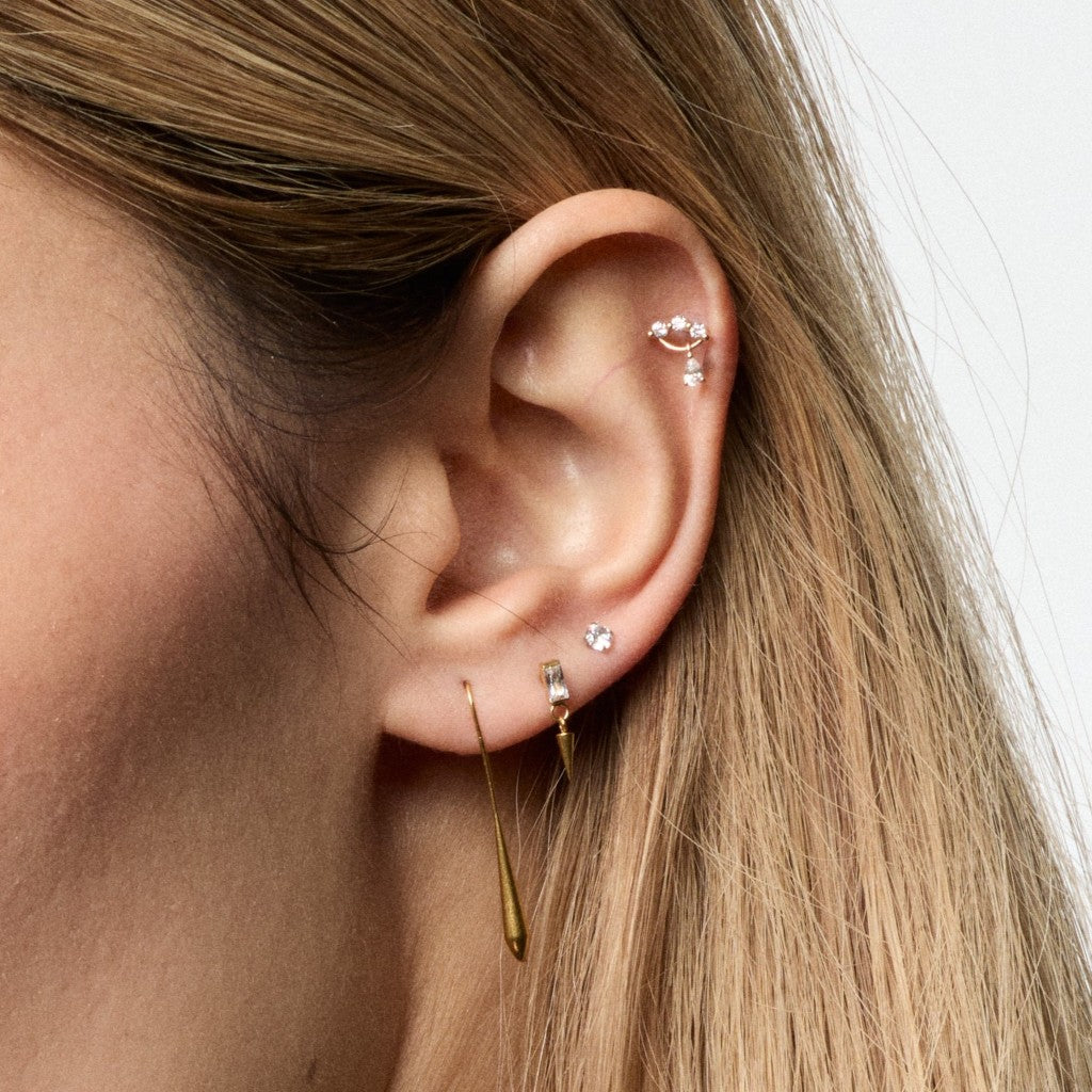 Close-up of a person's ear showcasing multiple gold and silver ear piercings, including a hoop in the cartilage, a small stud, and a long dangling teardrop earring, with blonde hair in the foreground against a white background.
