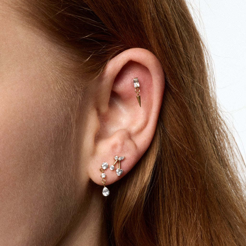 A close-up of a person's ear showcasing multiple gold-toned ear piercings: a small hoop in the lobe, a gold spike, and a cluster of clear gemstone studs with a dangling teardrop crystal.