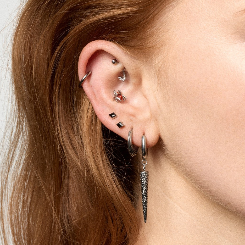 Close-up of a person's ear showcasing multiple piercings, including a silver cone dangle, silver hoop, and several small studs and cartilage piercings.