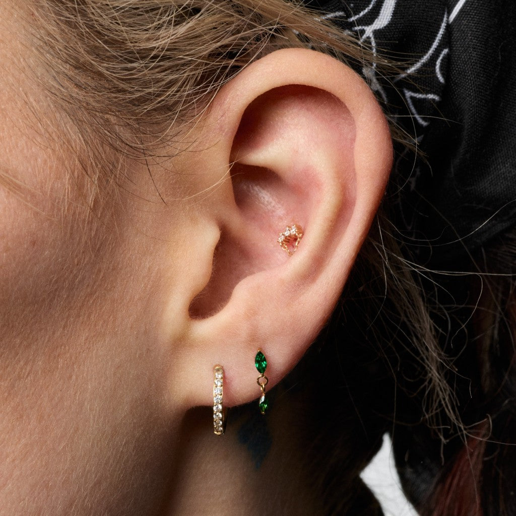 Close-up of a person's ear showcasing multiple small gold ear piercings, including a hoop earring with clear stones, a gold stud, and a green marquise-shaped gemstone earring.