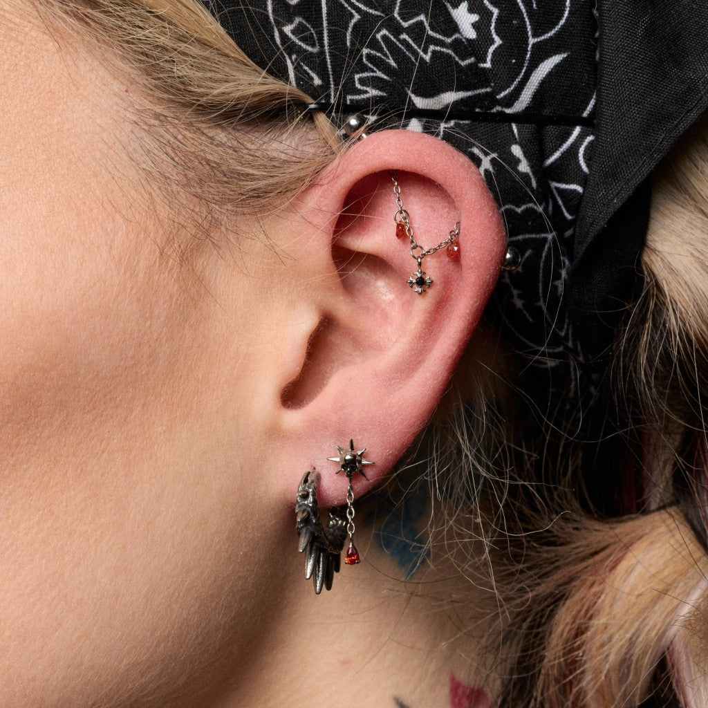 Close-up of a person's ear showcasing multiple gothic piercings, including a silver star-shaped cartilage piercing, a silver winged hoop in the lobe, and a silver ear cuff with red bead details.
