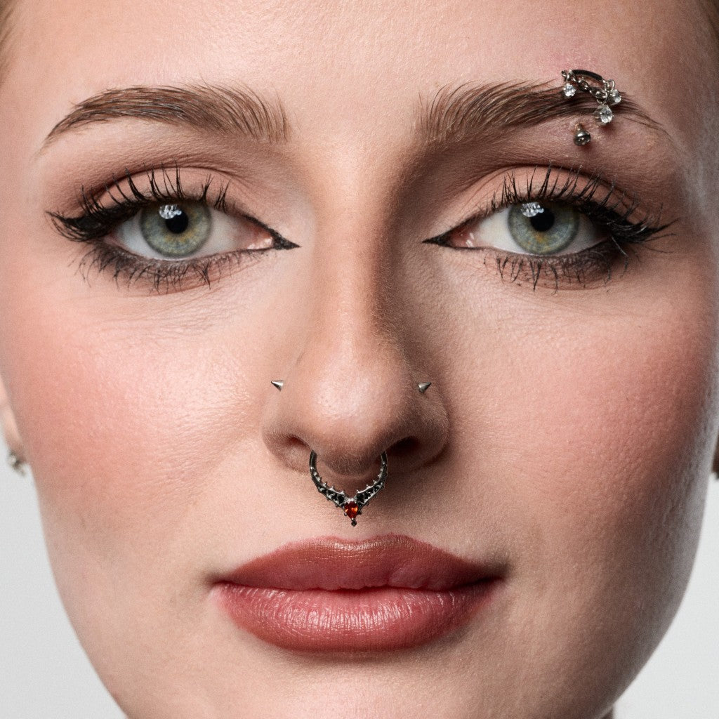 Close-up of a woman's face showcasing multiple facial piercings, including a silver nose hoop with red gemstones, a nose stud, and a brow piercing