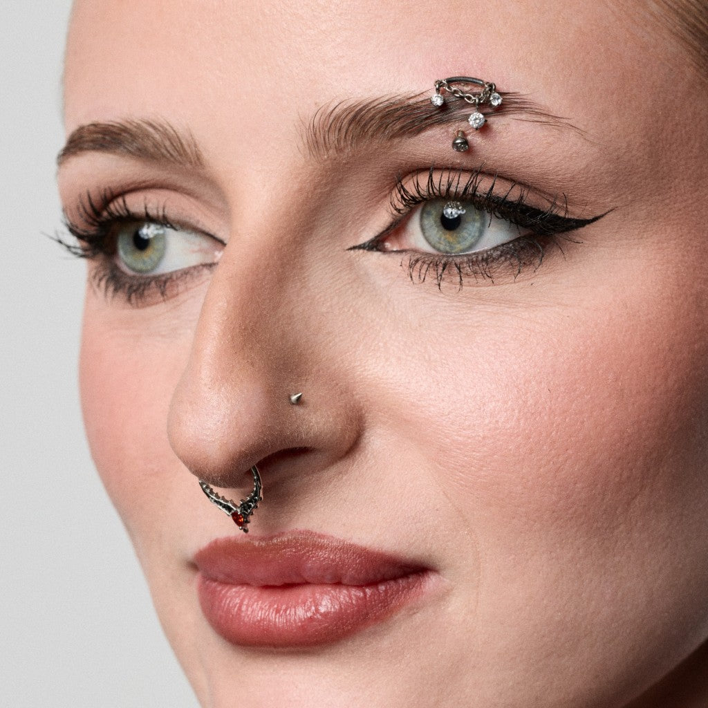 Close-up of a model's face showcasing multiple facial piercings: a silver eyebrow barbell with clear rhinestones, and a silver nose ring with dark red stones