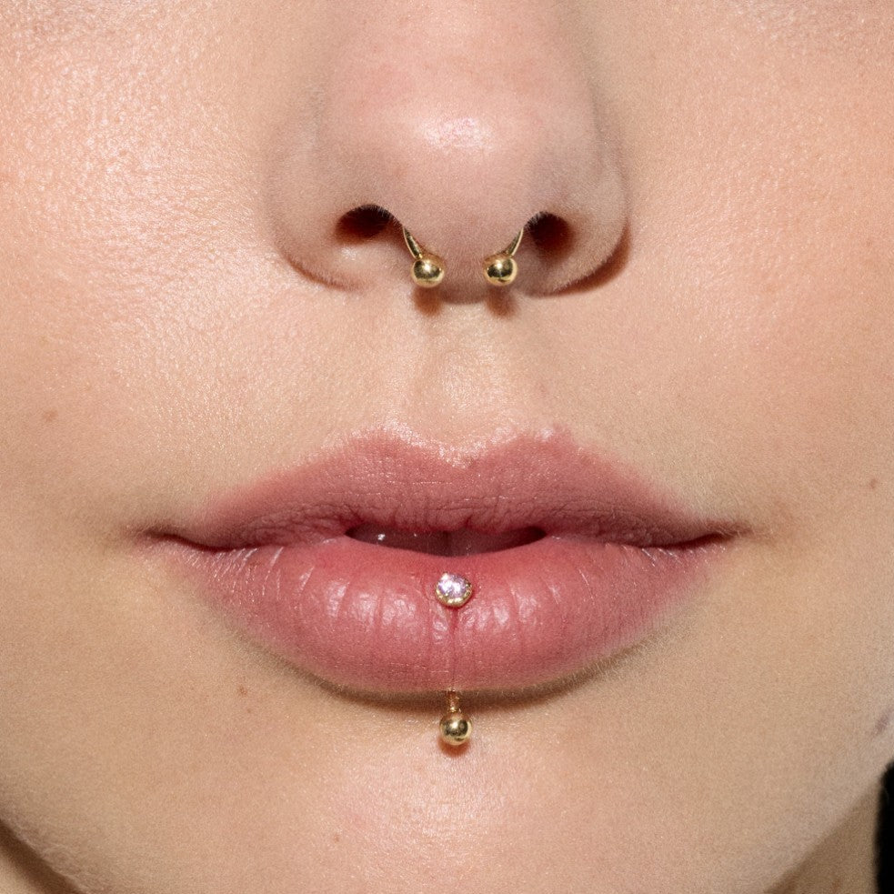 Close-up of gold septum ring and vertical labret piercing with clear cubic zirconia accent and polished gold ball ends.