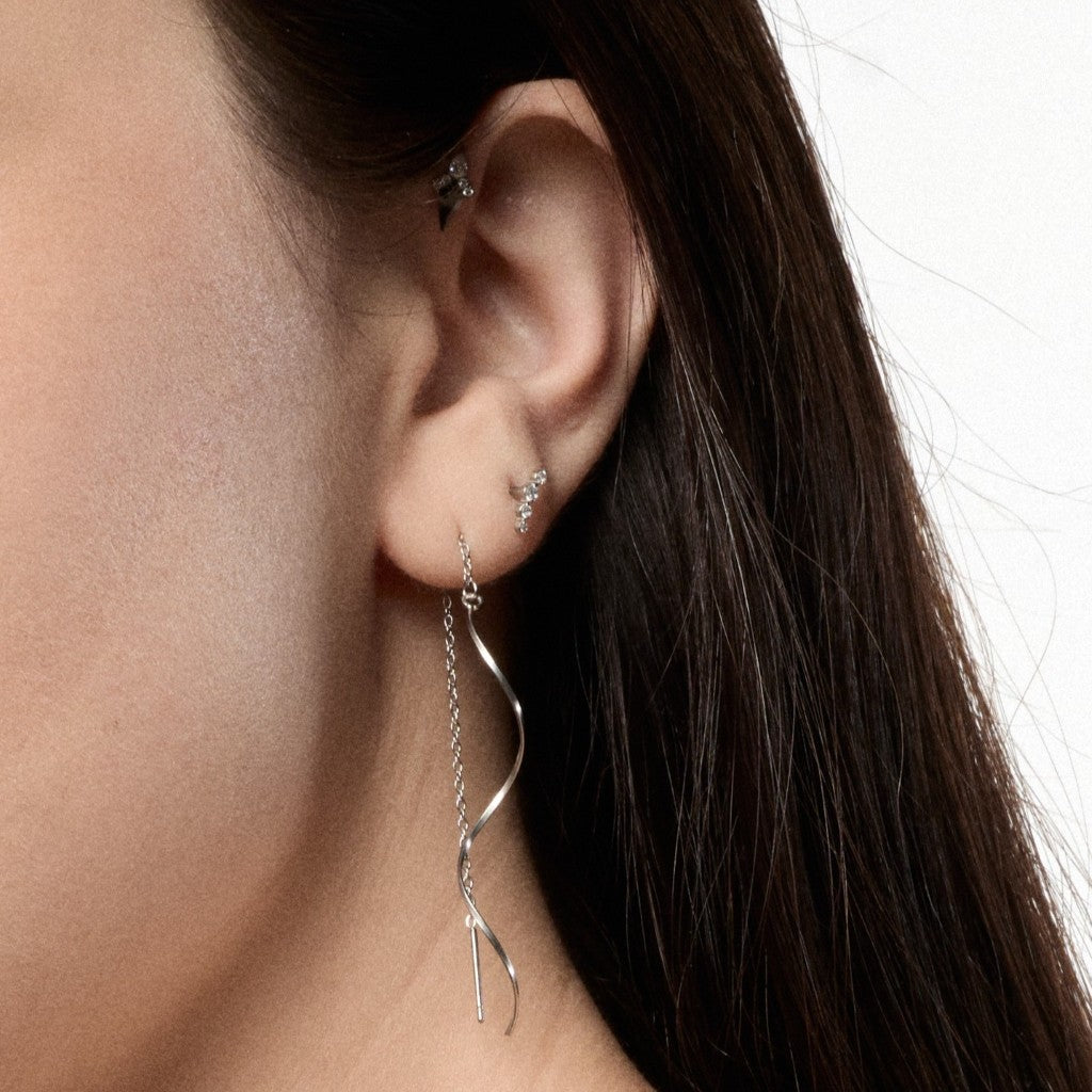 A close-up of a model's ear showcasing a silver dangle hoop earring with a twisted, snake-like chain that drapes down past the ear lobe. The model also has small stud earrings in other ear cartilage.